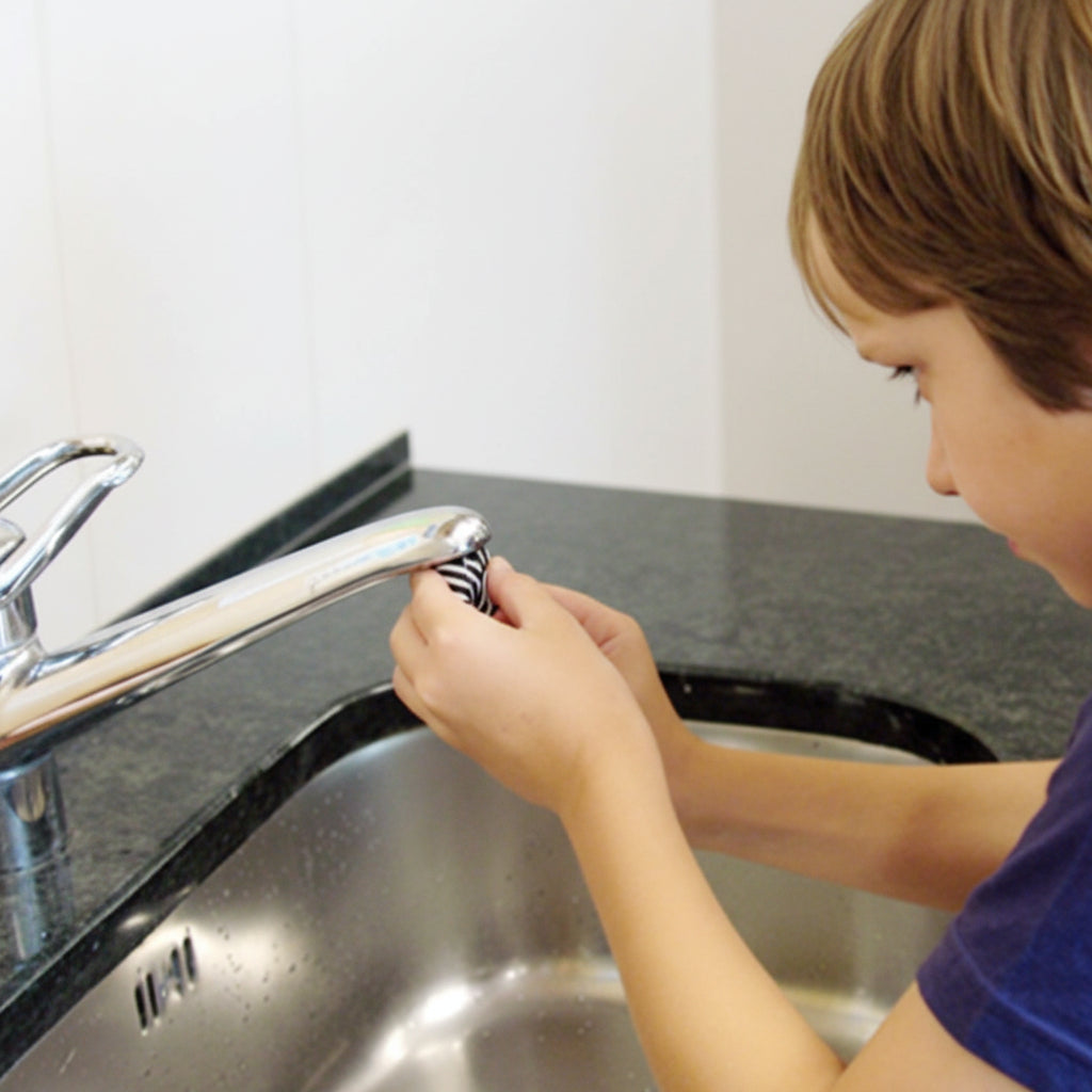 AquaClic Strahlregler Zebra Design Schwarz Weiss wird von einem kleinen Jungen ohne Werkzeug von Hand am Küchenwasserhahn über der Edelstahlspüle montiert
