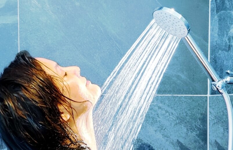 Attraktive Frau duscht mit geschlossenen Augten unter AquaClic-Duschkopf Prosecco, voller Wasserstrahl über Gesicht und hals; blau-grüne Fliesen im Hintergrund