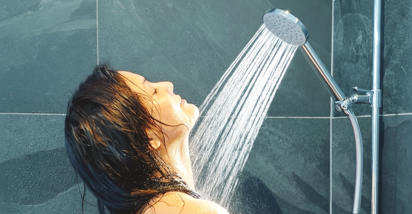 Unter wassersparender Duschbrause «Prosecco» an einer Duschstange duscht eine attraktive junge Frau, im Profil zu sehen mit geschlossenen Augen. Aus dem Duschkopf strömt ein voller Strahl füllig über Gesicht und Körper. Im Hintergrund blau-grüne Steinkacheln.